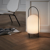 Neutral white lamp with ribbed pattern, supported by a sleek black frame, set against warm wooden flooring.