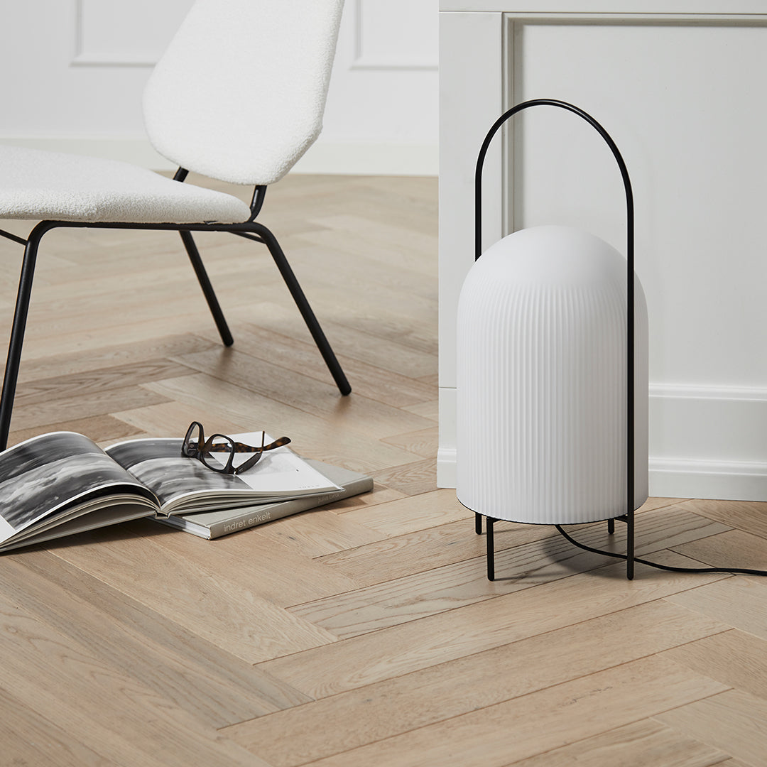 Minimalist white lamp with ribbed texture and sleek black stand, set against warm wooden herringbone floor.