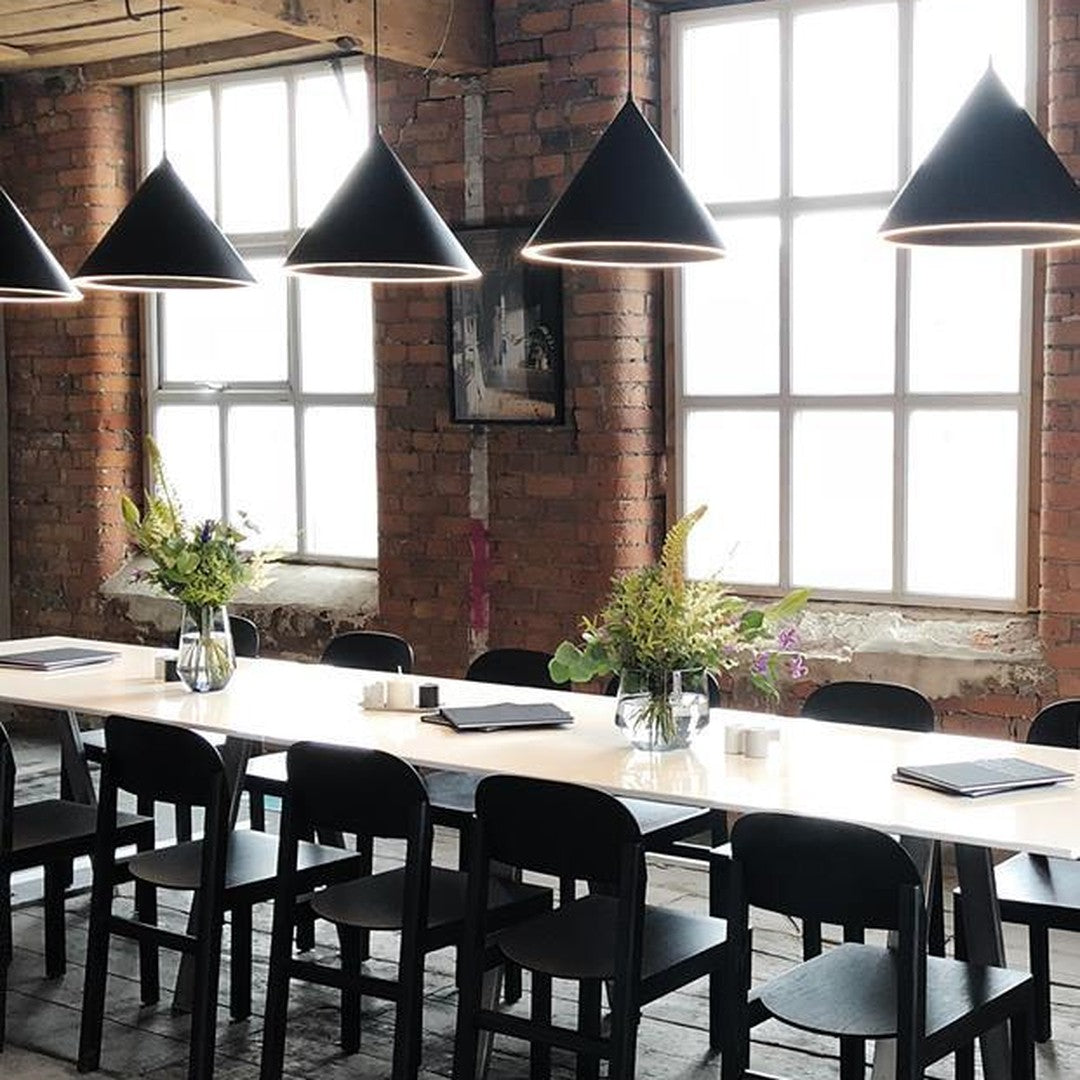 Modern dining setup featuring black cone pendant lights, warm brick walls, and a sleek white table. Green floral accents.