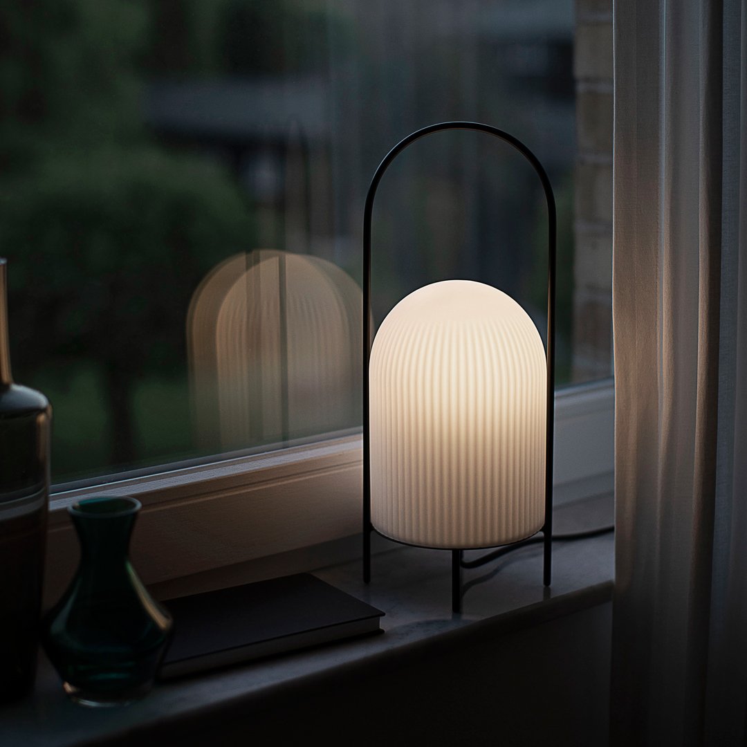 Minimalist black lamp with a ribbed white shade, illuminating a warm, soft glow against a green backdrop.