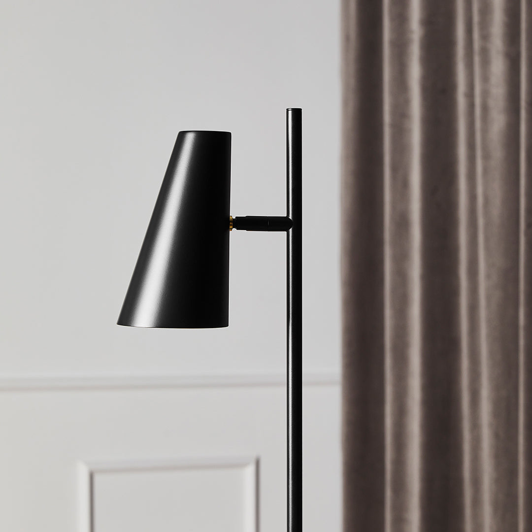 Black modern lamp with a sleek cone shade against a soft gray backdrop and elegant curtains.