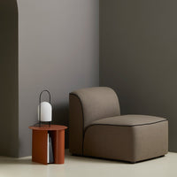 Curved, taupe armchair with contrasting dark trim against a gray wall and a warm terracotta side table. Soft, minimalist design.