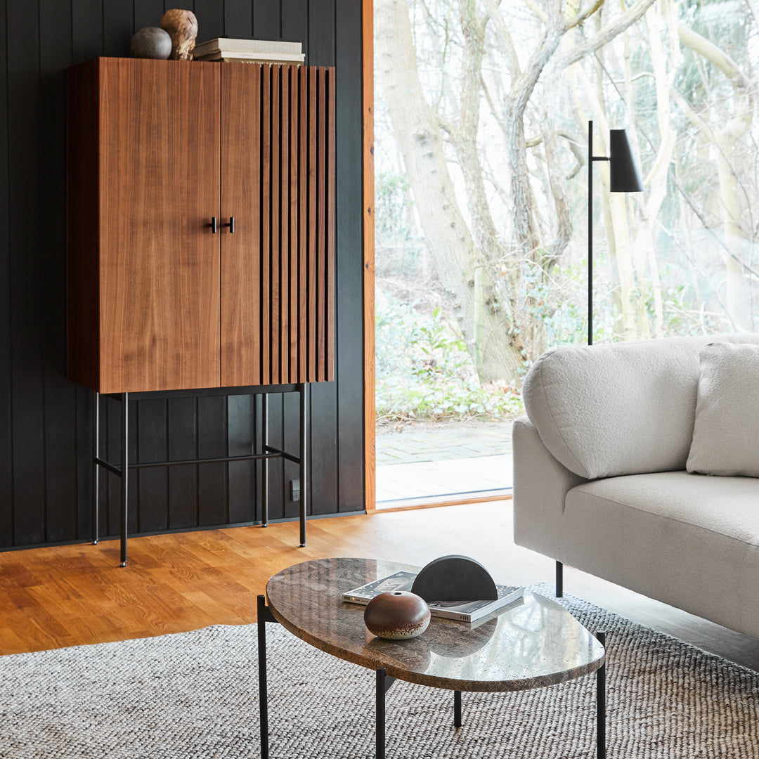 Wooden cabinet with vertical slats, sleek black legs, warm wooden floor, and neutral-toned furniture in a modern setting.