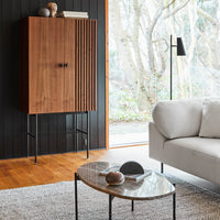 Wooden cabinet with vertical slats, sleek black legs, warm wooden floor, and neutral-toned furniture in a modern setting.