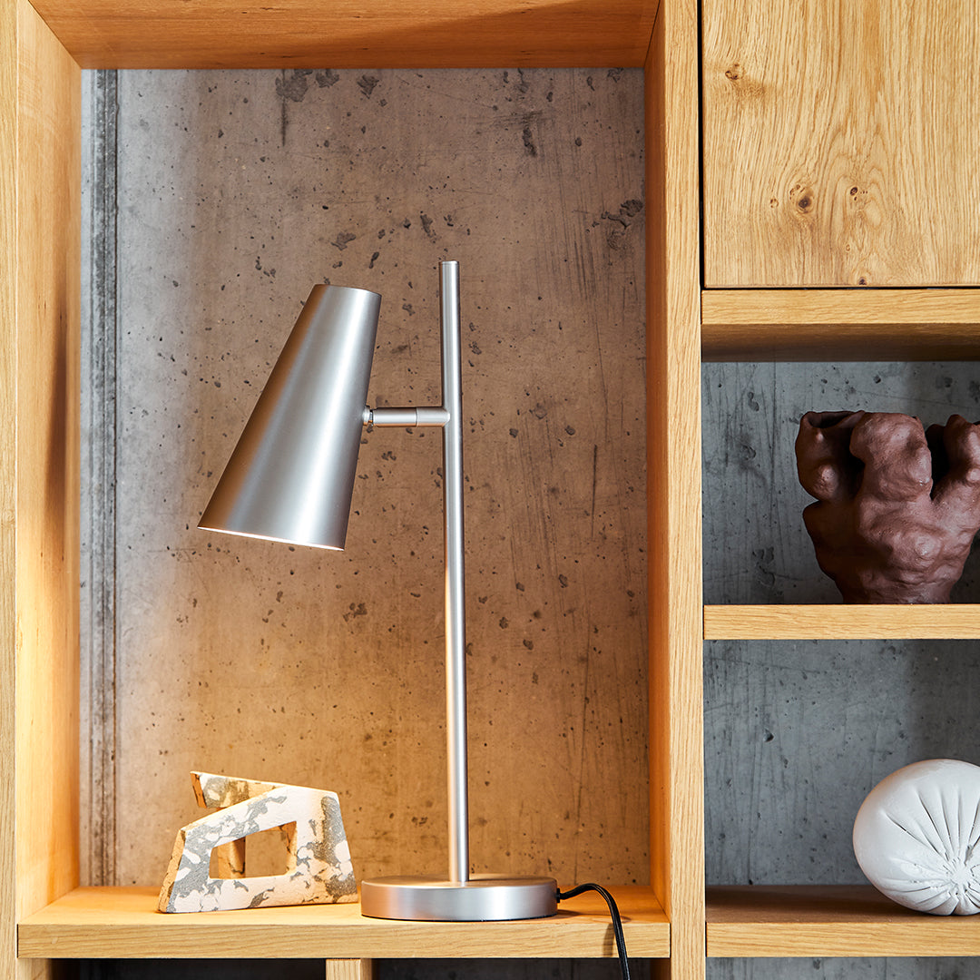 Sleek silver desk lamp against a textured concrete backdrop, surrounded by warm wooden shelves.