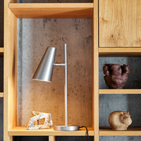 Silver desk lamp with a sleek design, displayed against warm wooden shelves and a concrete backdrop.