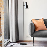 Black metal standing lamp beside a gray sofa with a textured orange cushion and soft curtains in a light room.