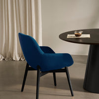 "Vibrant blue upholstered chair with smooth contours against a neutral backdrop and dark round table."