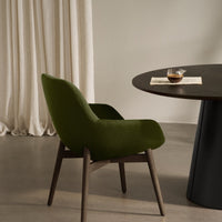 Olive green upholstered chair with wooden legs beside a dark round table, complemented by beige curtains in the background.