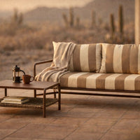 Beige and cream striped outdoor sofa with a matching throw, paired with a wooden coffee table in a desert setting.