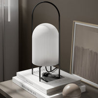 White ribbed glass lamp with a sleek black frame, set on stack of neutral-toned books on a soft gray surface.