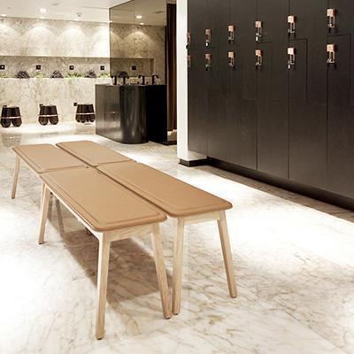 "Light tan upholstered bench with natural wood legs, set against a sleek marble floor and dark locker backdrop."