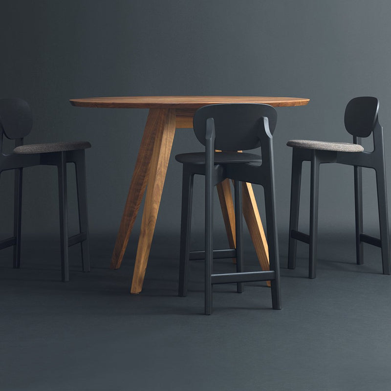 "Round wooden table with natural finish and sleek black bar stools against a dark gray backdrop."