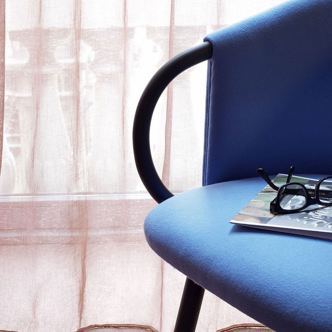 Blue chair with smooth fabric, black metal frame, and a backdrop of soft, sheer pink curtains.