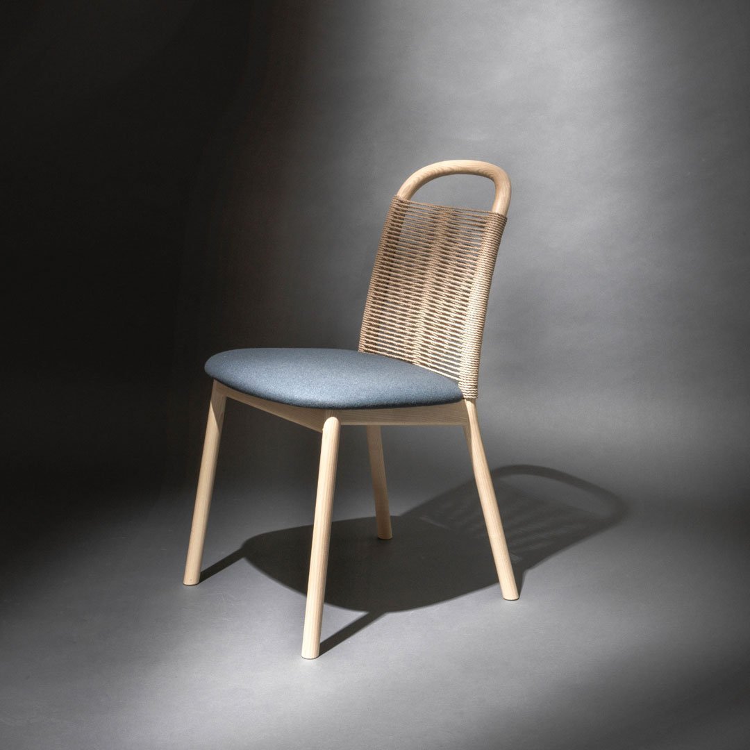 Light wood chair with a woven back and blue cushion, featuring a modern, minimalist design against a dark backdrop.