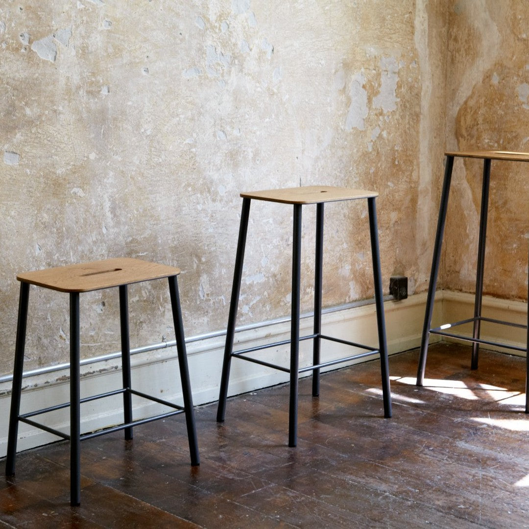 "Three minimalist bar stools with natural wood tops and black metal legs against a textured beige wall."