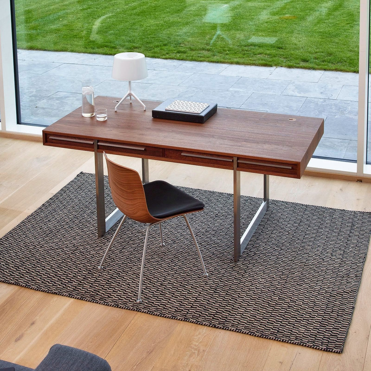 Modern wooden desk in rich brown with a sleek metal base, paired with a black chair on a textured gray rug.