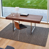 Modern wooden desk in rich brown with a sleek metal base, paired with a black chair on a textured gray rug.