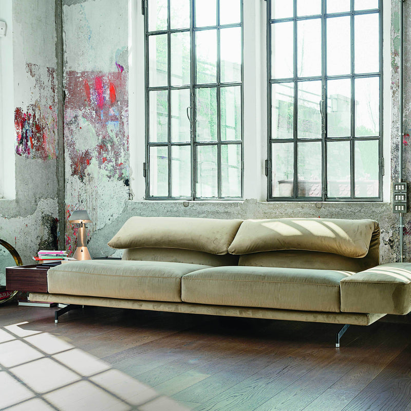 Beige modern sofa with sleek lines, set against an industrial backdrop of textured walls and large windows.