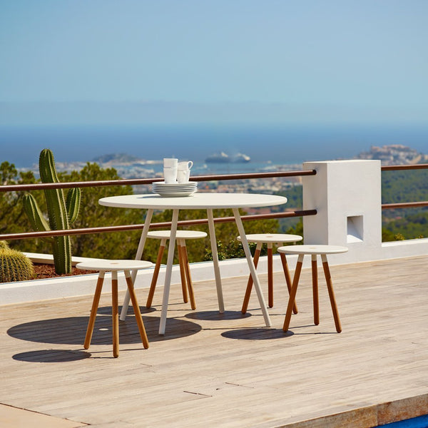 Stylish white outdoor table with natural wood legs, set against a serene blue ocean view and lush green plants.