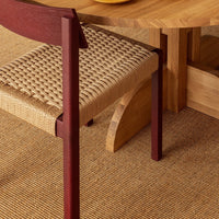 Dark red chair with woven beige seat, paired with a light wooden table and textured tan rug beneath.