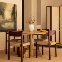 Round wooden dining table with maroon chairs, beige upholstery, minimalist design, and natural wood tones.