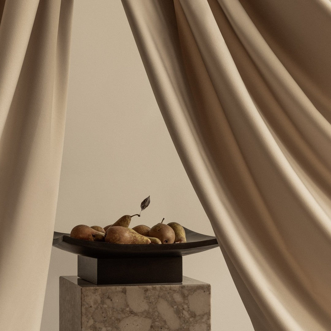 Beige curtains frame a dark plate with pears, set on a light marble pedestal, creating a soft, elegant aesthetic.