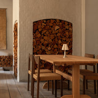 Light wood dining table with natural wicker chairs, complemented by textured beige walls and stacked brown wood backdrop.
