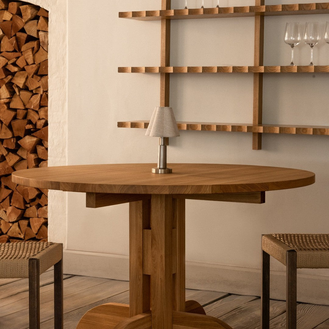 Round wooden dining table with a warm brown finish, featuring a smooth surface and simple design. Neutral tones and natural textures.