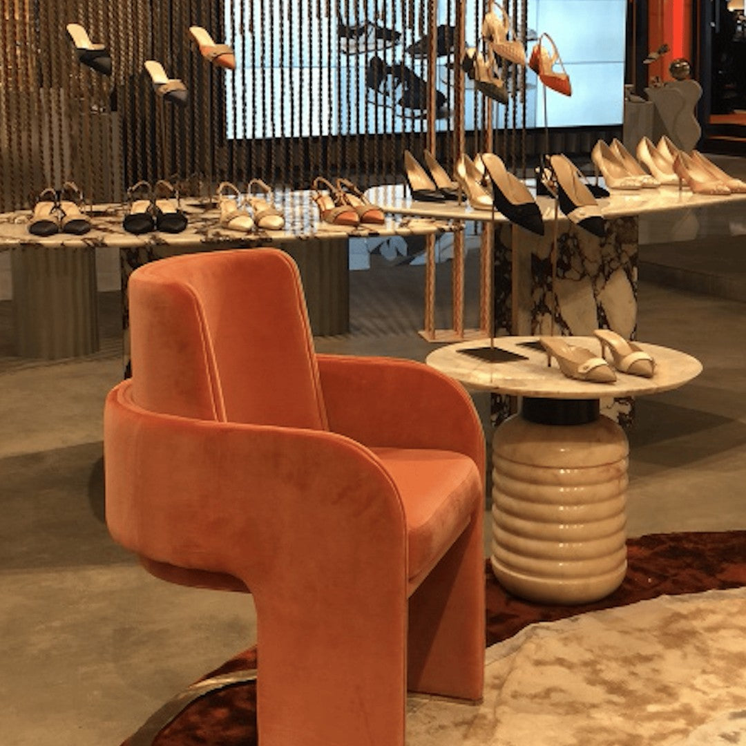 Velvet pink chair with curved design, set against a neutral-toned floor and stylish shoe display backdrop.