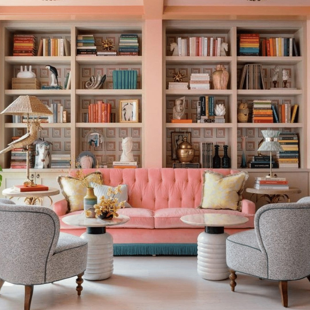 Colorful living room with a pink tufted sofa, gray patterned chairs, and vibrant bookcases featuring bold hues and pastel accents.