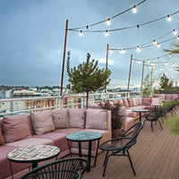 "Soft pink cushions and patterned tables against a serene skyline backdrop with warm string lights."