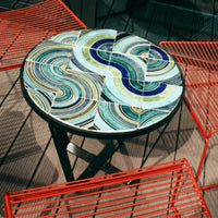 Colorful mosaic table featuring swirling green, blue, and white patterns with a sleek black frame and vibrant red chairs.