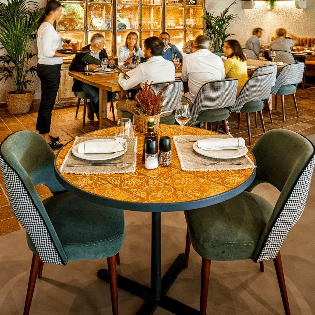 Round table with intricate wood pattern, green velvet chairs, and neutral-toned decor in a lively dining setting.