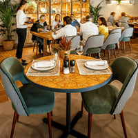 Round table with intricate wood pattern, green velvet chairs, and neutral-toned decor in a lively dining setting.