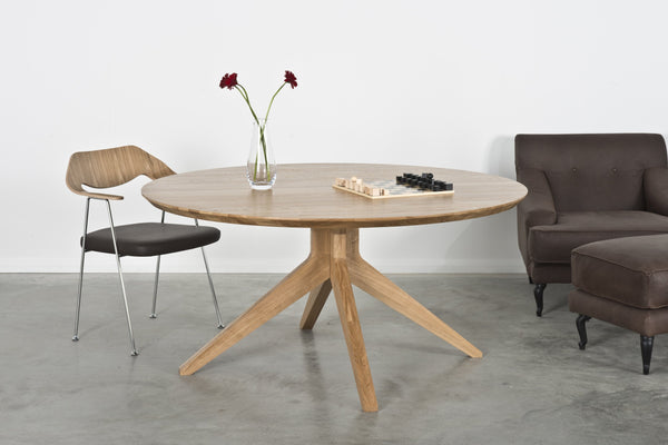 Light wood round dining table with a smooth surface and tapered legs, paired with a neutral-toned chair and dark couch.