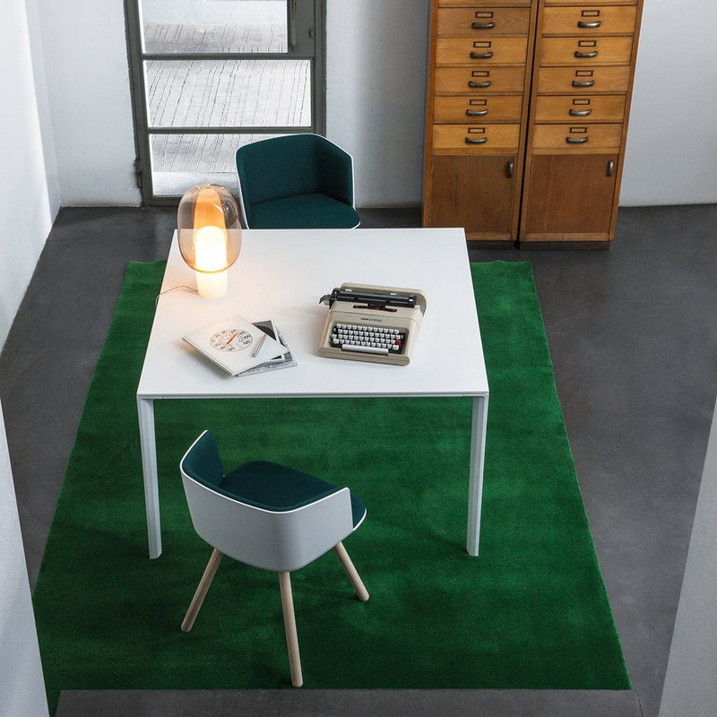 Modern workspace featuring a white table, teal chair, and vibrant green rug in a stylish, minimalist setting.