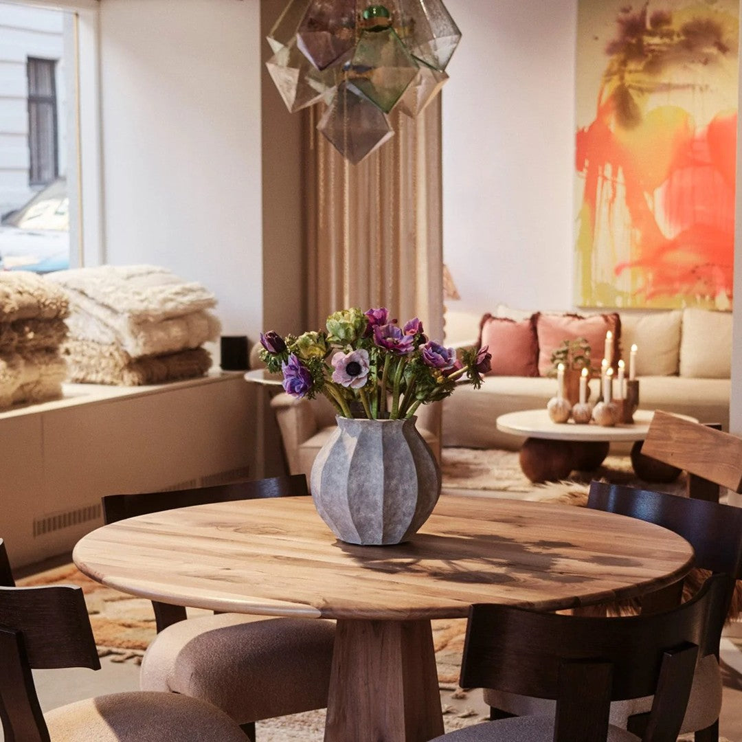 Wooden table with a geometric vase, filled with purple flowers, surrounded by neutral tones and soft textures in the room.