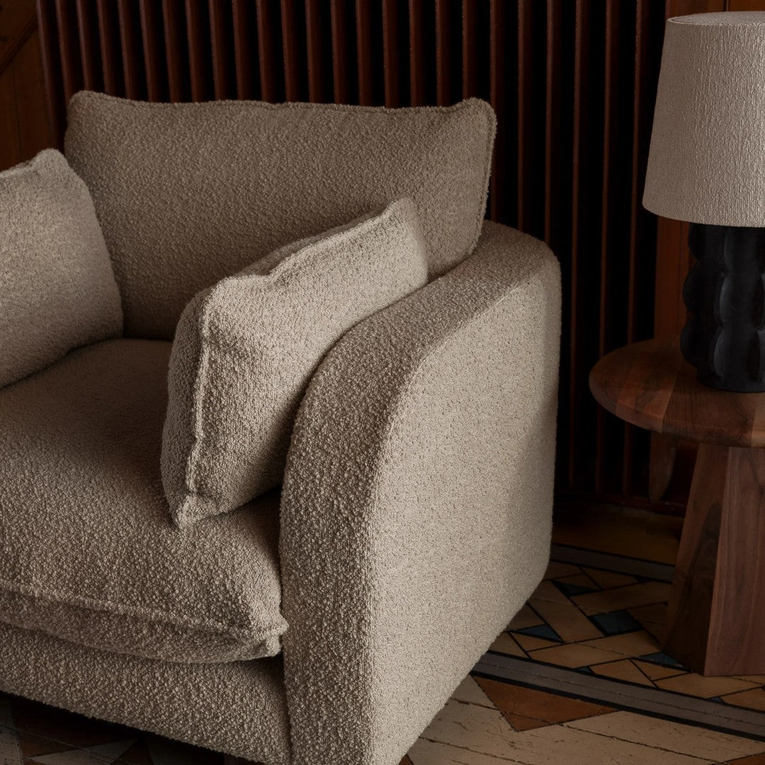Beige textured armchair with plush cushions and soft, rounded edges, paired with a wooden side table and lamp.