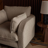 Beige textured armchair with plush cushions and soft, rounded edges, paired with a wooden side table and lamp.