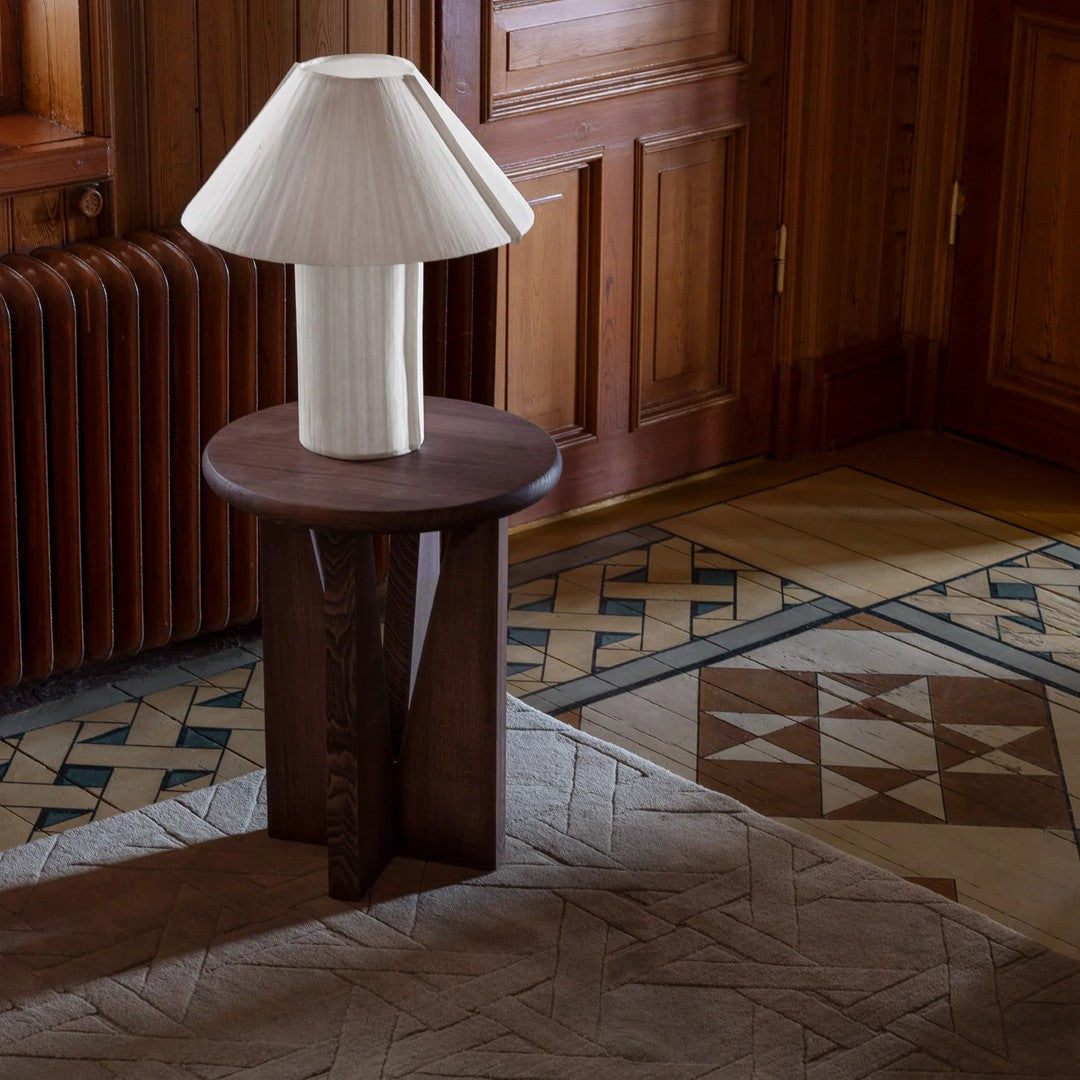 Elegant white pleated lamp on a rustic brown wooden table, set against geometric patterned flooring in neutral tones.