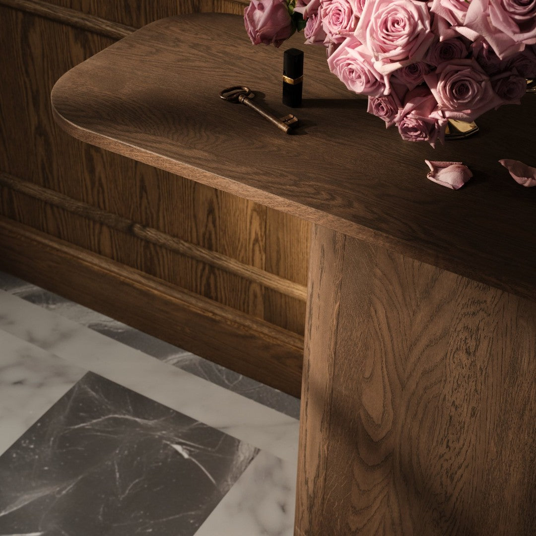 Close-up of a warm wood table with soft curves, alongside a bouquet of pink roses and a marble patterned floor.