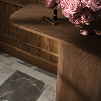 Close-up of a warm wood table with soft curves, alongside a bouquet of pink roses and a marble patterned floor.