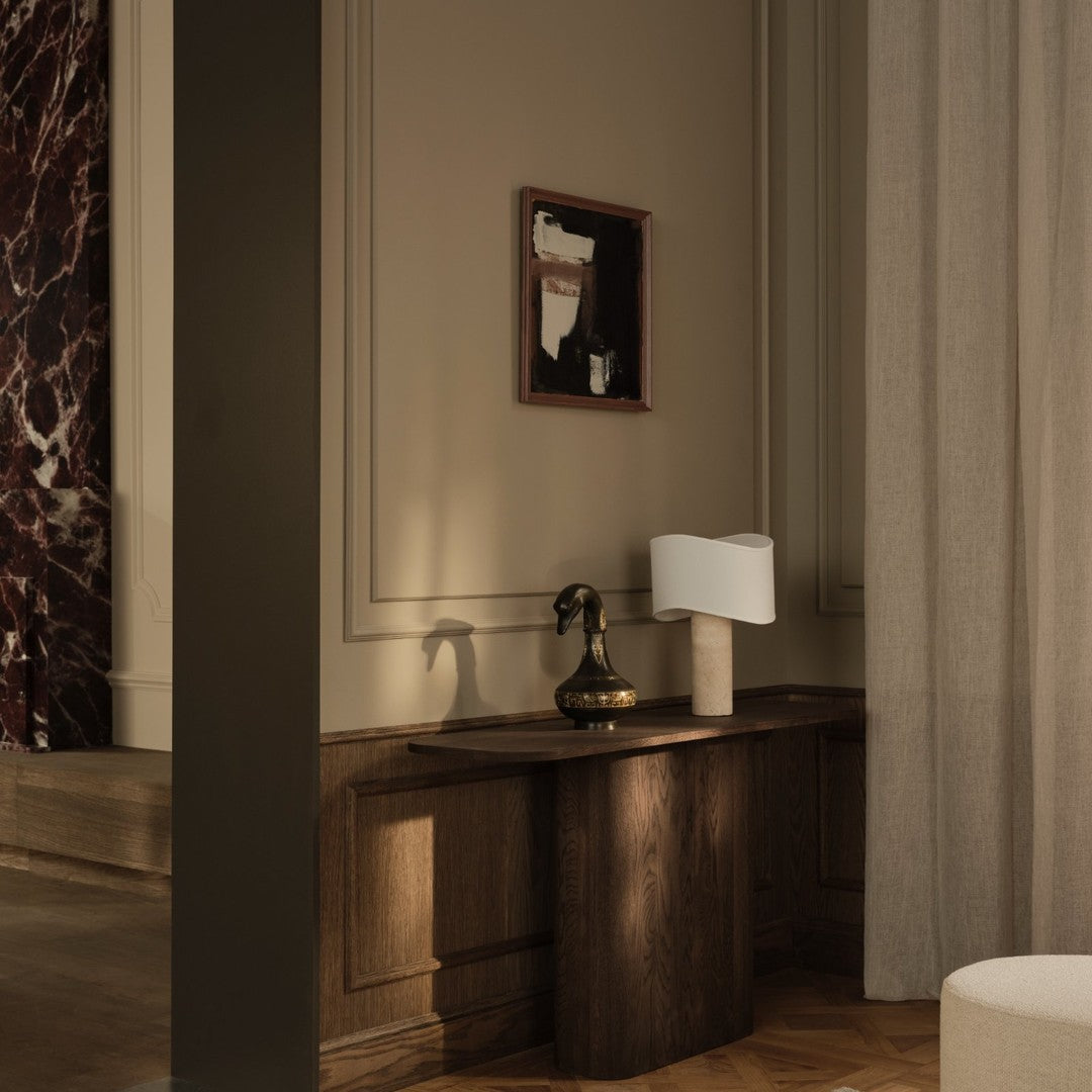 Contemporary wooden table with a sleek white lamp, dark wall art, and soft gray curtains in a warm-toned room.