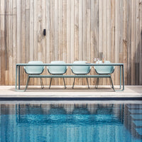 Mint green table and chairs against a natural wood backdrop, with a tranquil blue pool reflecting the surroundings.