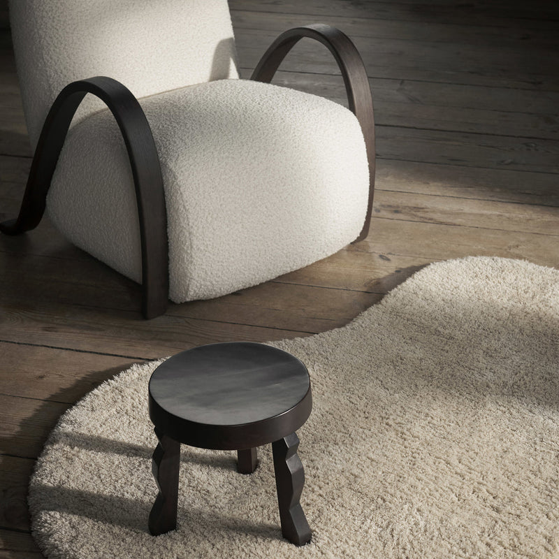 Cozy cream rug with a wavy pattern, accompanied by a round black wooden stool and a neutral upholstered chair.