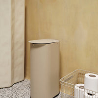 Beige cylindrical trash can against a soft yellow wall, complemented by a textured gray terrazzo floor.