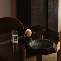 Black textured bowl on a glass table, accompanied by a clear glass and yellow apple, with dark brown chairs in the background.