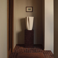 Elegant white vase on dark pedestal, contrasted against warm beige walls and earthy red brick flooring.