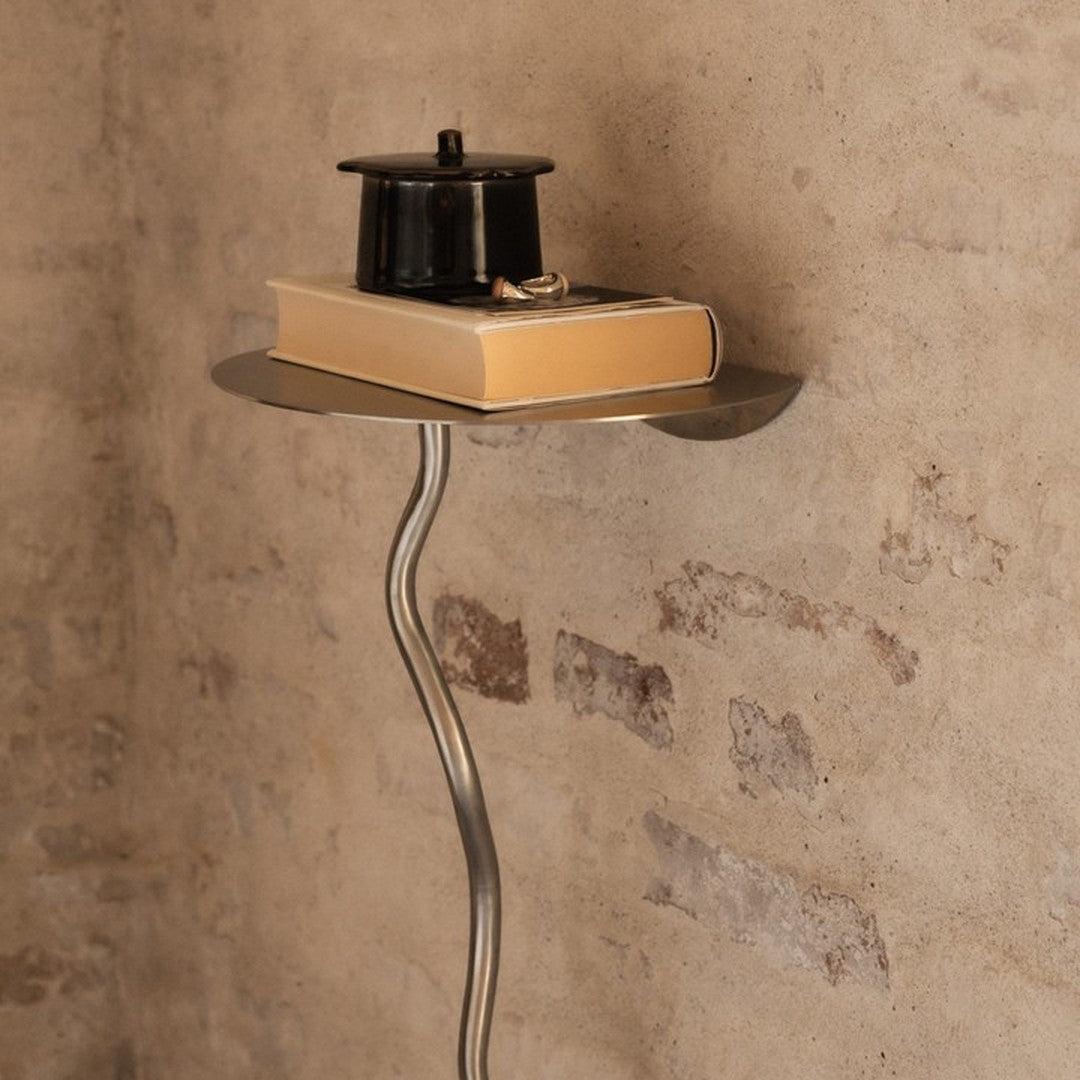 Stylish silver metal stand with a smooth, circular top, displaying a black container and a beige book against a textured wall.